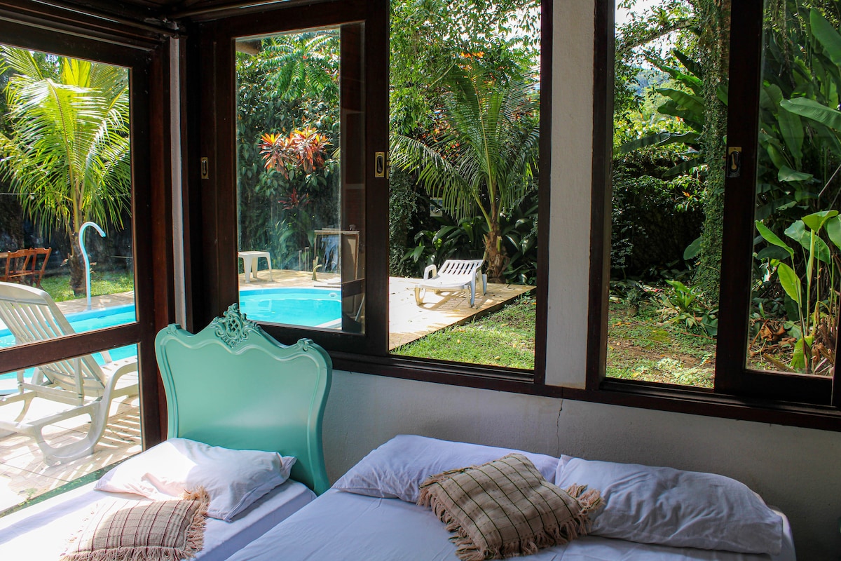 A bright sleeping area features two beds with soft linens and decorative pillows. Large windows frame a view of the lush garden and shared pool in the outdoor space, inviting natural light into the room.