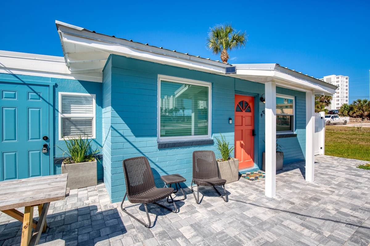 A vibrant exterior features a blue façade accented by a coral front door. Two dark chairs are arranged on the patio, inviting relaxation. The surrounding area is paved with light-colored stones, and palm trees are visible against a clear blue sky.