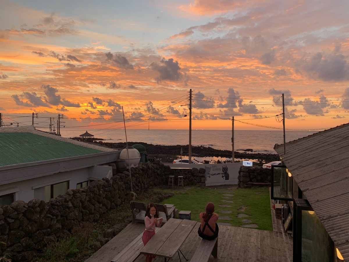 The image captures a vibrant sunset over the ocean, with hues of orange and pink filling the sky. In the foreground, a wooden table sits among a grassy area, while guests are seen enjoying the view. The rustic stone wall adds texture to the setting.