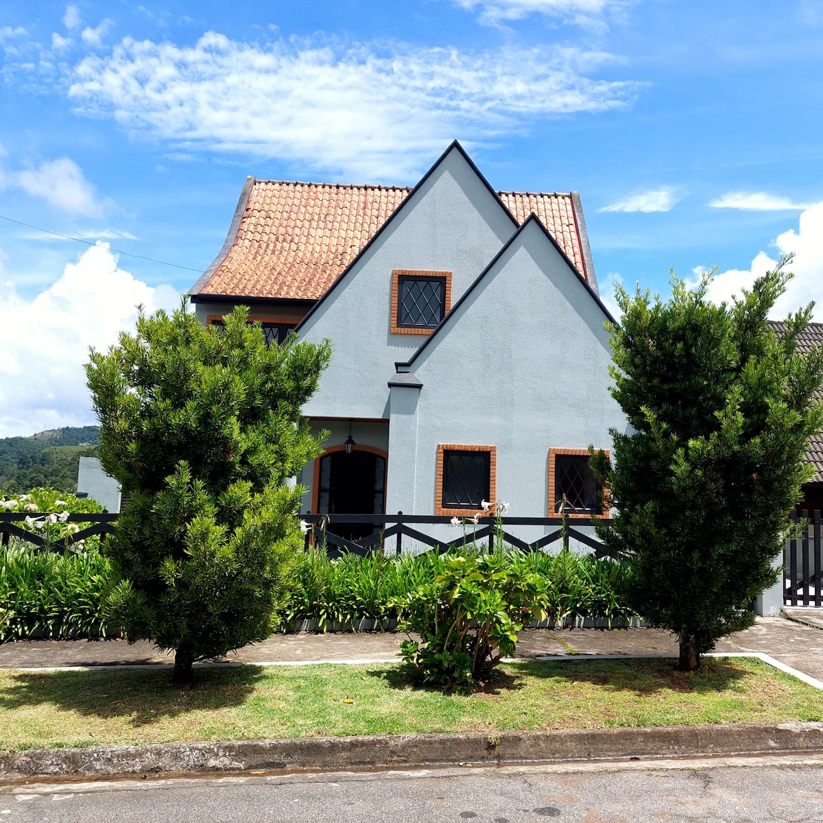 A charming three-story house is set against a bright sky, showcasing a distinct architectural style with a tiled roof and angled eaves. Lush greenery lines the yard, with well-maintained shrubs framing the entrance and providing a welcoming appearance.