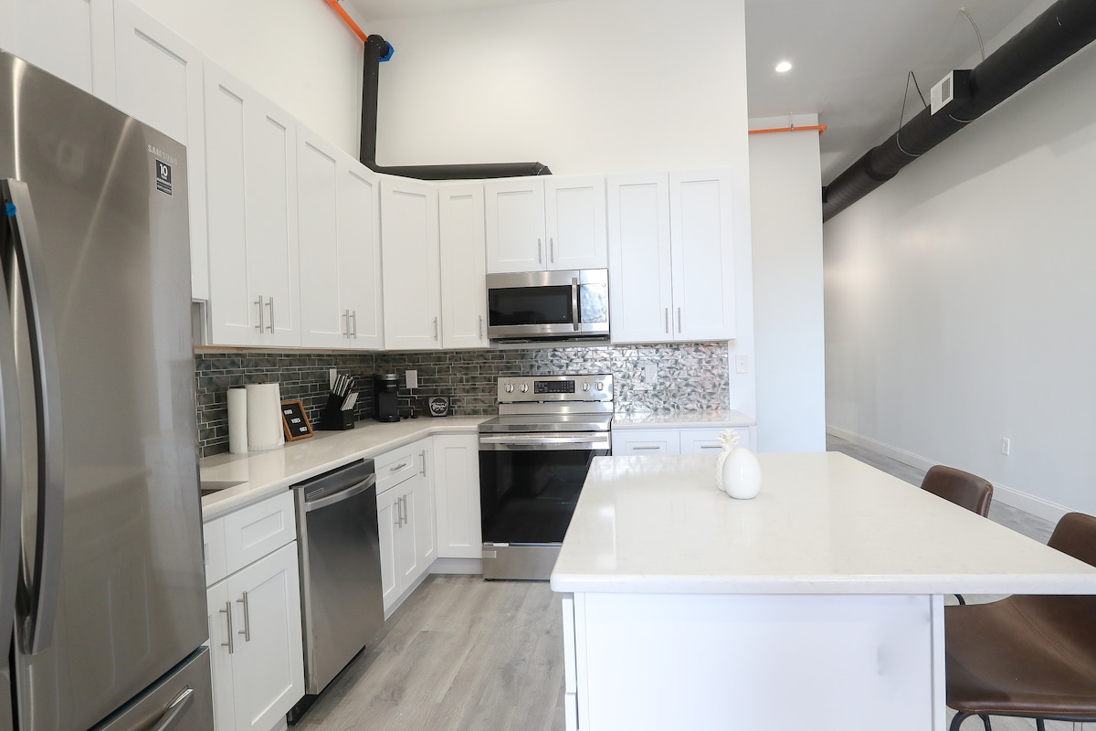 The modern kitchen features white cabinetry, stainless steel appliances, and a light-colored stone countertop. A mosaic backsplash adds texture, while a central island provides additional preparation space. Several high-backed chairs are positioned at the island, complementing the bright and airy atmosphere.