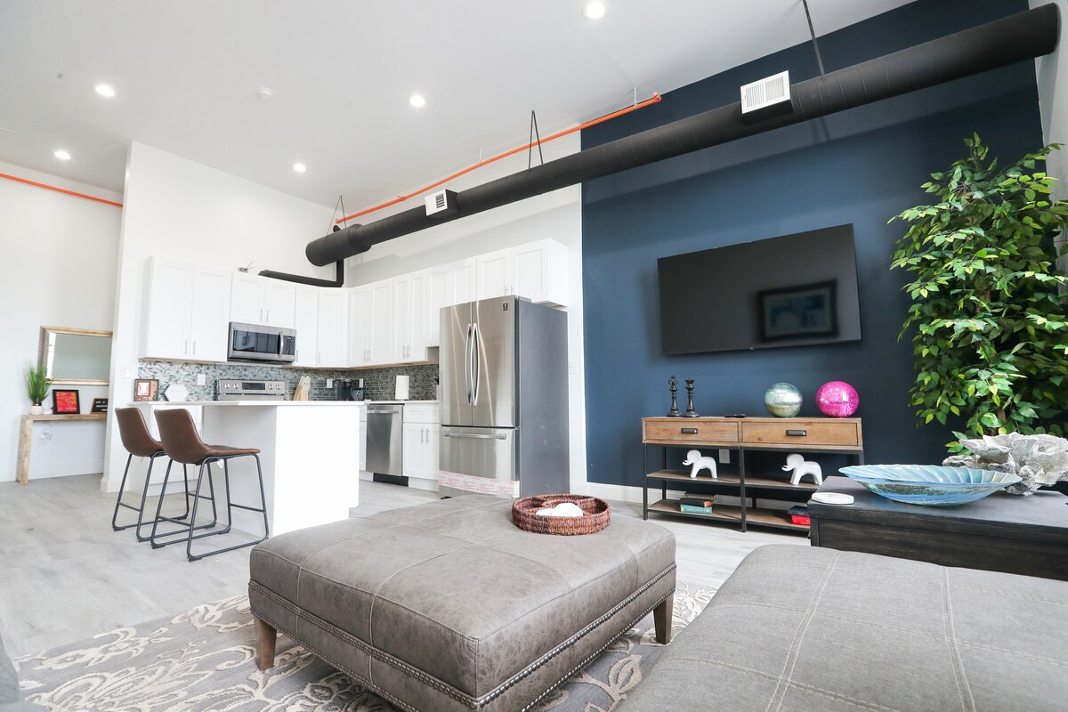 A spacious living area features a large ottoman in the foreground, with a modern kitchen area equipped with stainless steel appliances visible in the background. White cabinetry contrasts with a dark blue accent wall, while decorative plants add a touch of greenery to the scene.