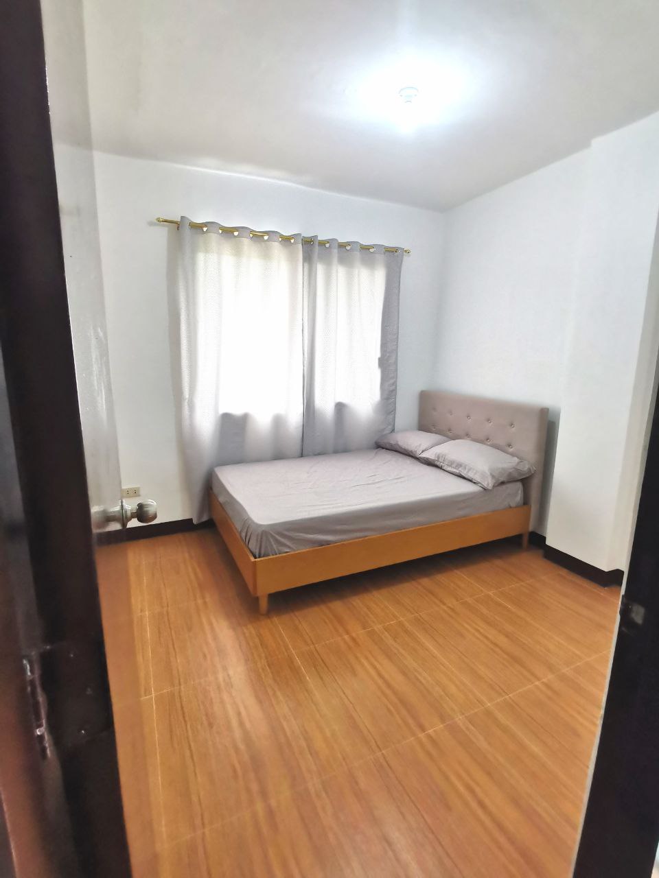 A simple bedroom features a comfortable bed with a light-colored headboard. Soft curtains frame the window, allowing natural light to brighten the space. Wooden flooring complements the muted color palette, creating a relaxed atmosphere.