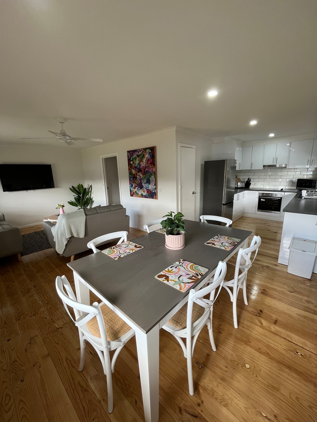 An open living space showcases a modern dining table with six white chairs, set with colorful placemats. In the background, a comfortable seating area is visible with a large smart TV and a ceiling fan above, creating a bright and welcoming atmosphere.