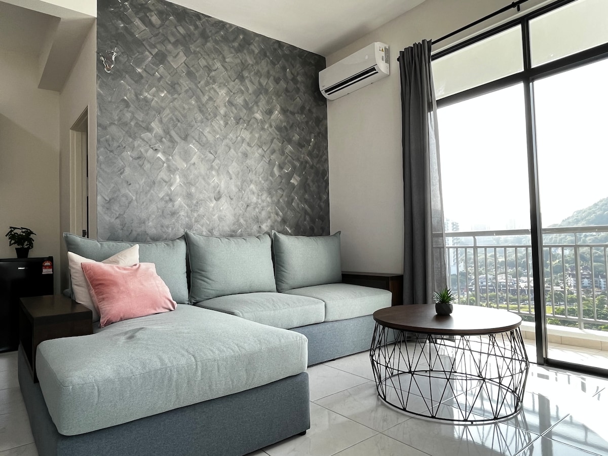 A spacious living area features a large, light blue sectional sofa with a pink accent pillow. A round, wire coffee table is positioned in front. Light filters in through a floor-to-ceiling window, revealing views of surrounding hills and a cityscape.