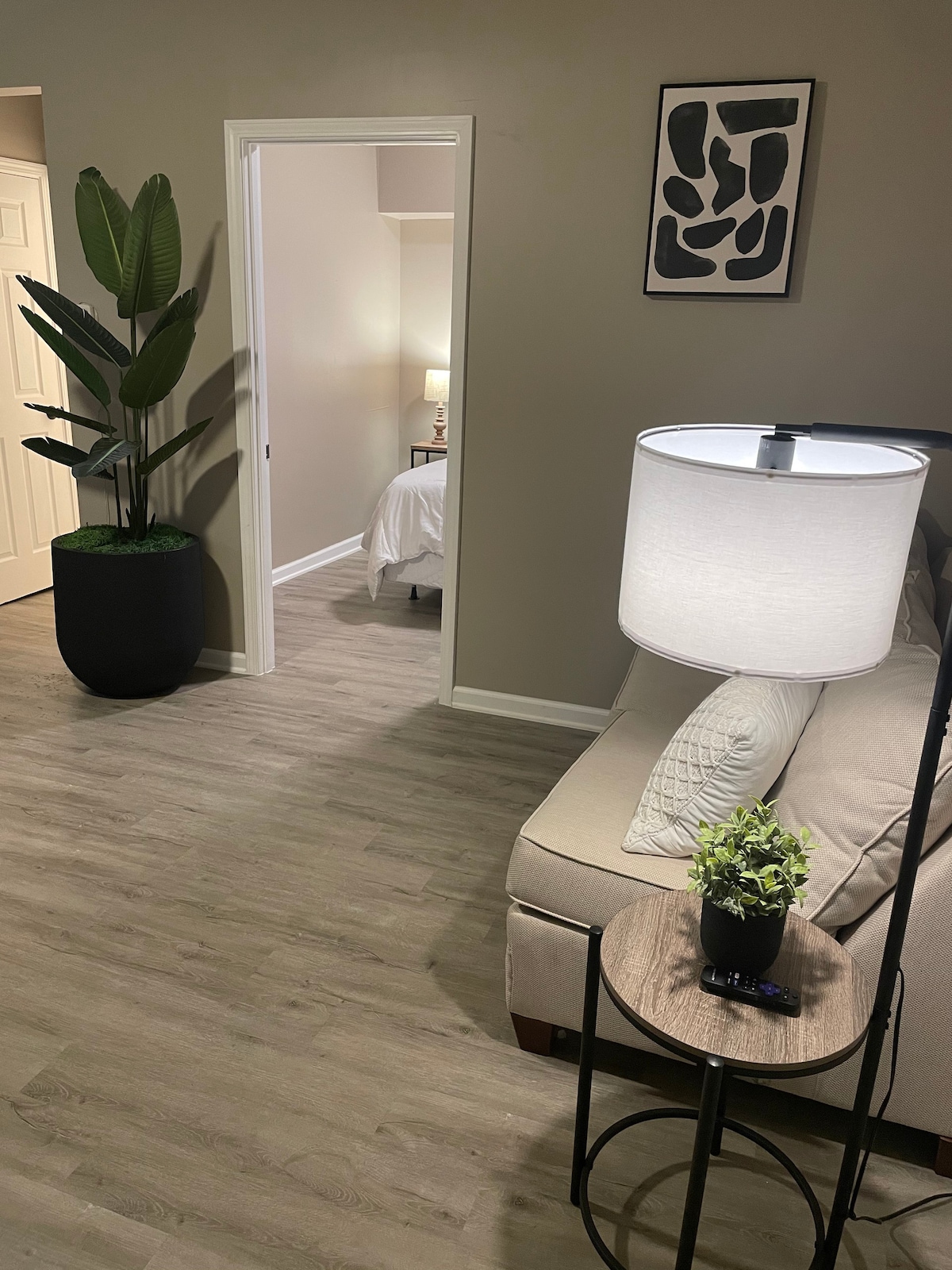 A contemporary living area is highlighted by a soft sofa and a round side table, topped with a small potted plant. A tall plant adds a touch of greenery, while a doorway leads to the bedroom area in the background, enhancing the spacious feel of the space.