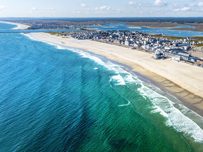 Aerial view captures the expansive shoreline of Hampton Beach, where gentle waves meet sandy shores. A clear blue ocean contrasts with the vibrant green waters closer to the beach, while the distant coastal town is visible against a backdrop of a bright sky.