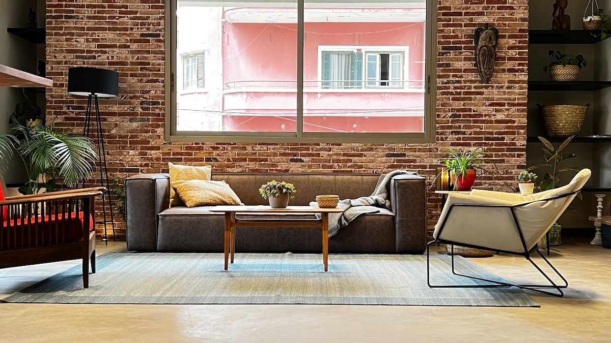 A spacious living area features a cozy sectional sofa, wooden coffee table, and stylish accent chair. The exposed brick wall adds character, while large windows allow natural light to fill the space. Decorative plants are placed strategically, enhancing the urban modern aesthetic.