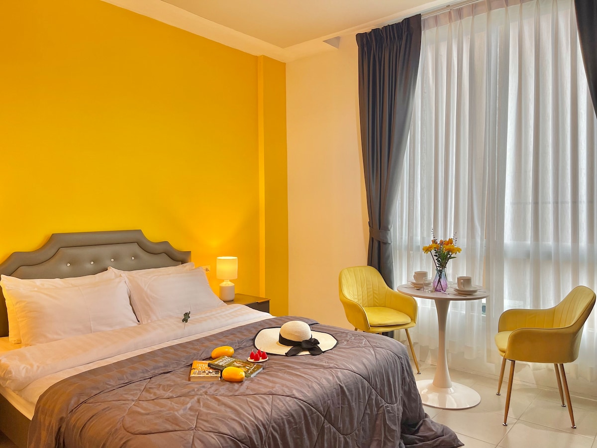 A bright studio room features a queen-size bed with a gray comforter, complemented by yellow walls. A small round table with two chairs occupies one corner, adorned with a vase of flowers. Natural light filters through sheer curtains, adding warmth to the space.