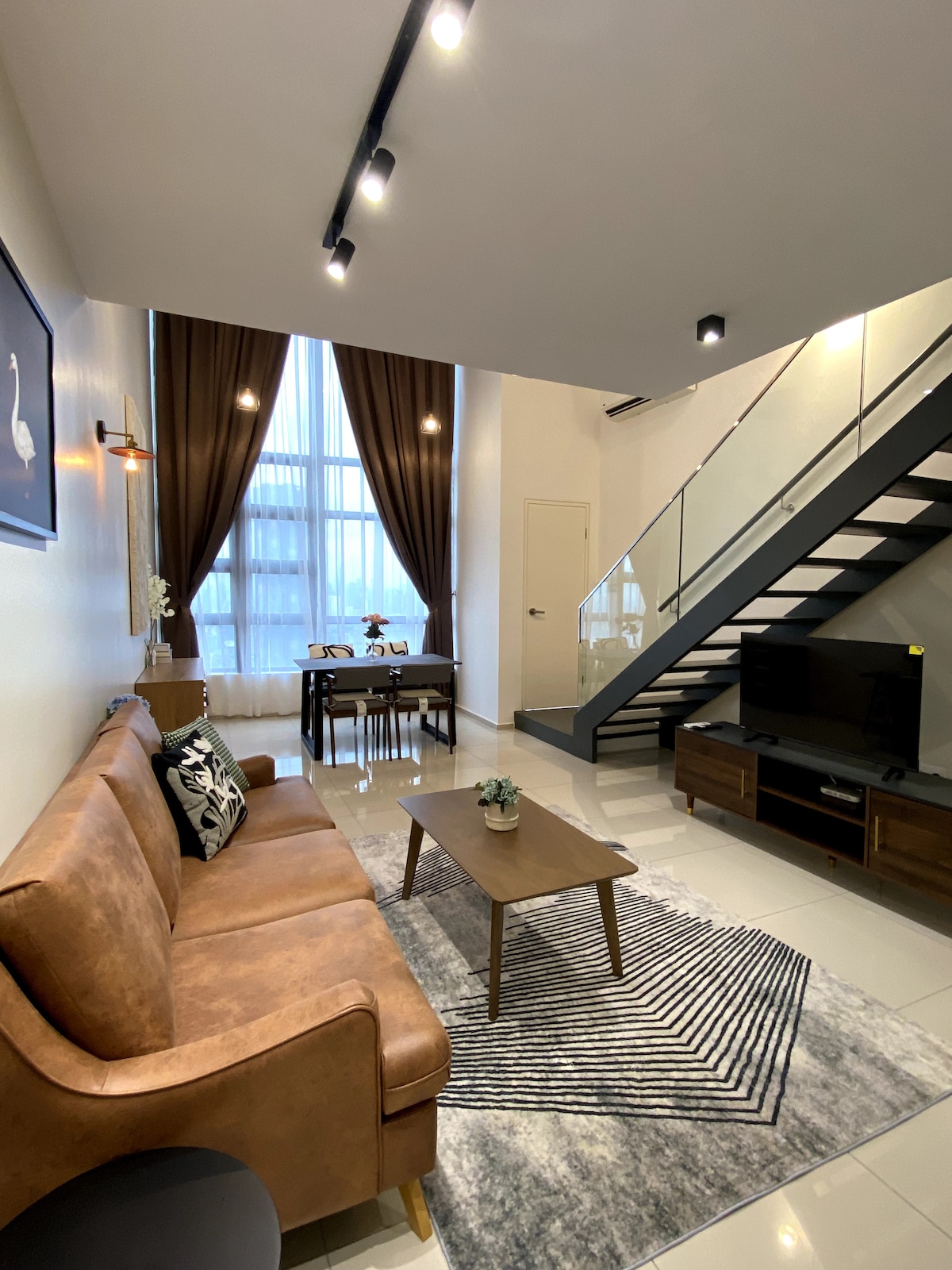 An airy living area is showcased with tall windows allowing natural light. A brown sofa and geometric rug are present, complemented by a small wooden coffee table. A dining table is visible in the background, with modern lighting fixtures enhancing the contemporary design.