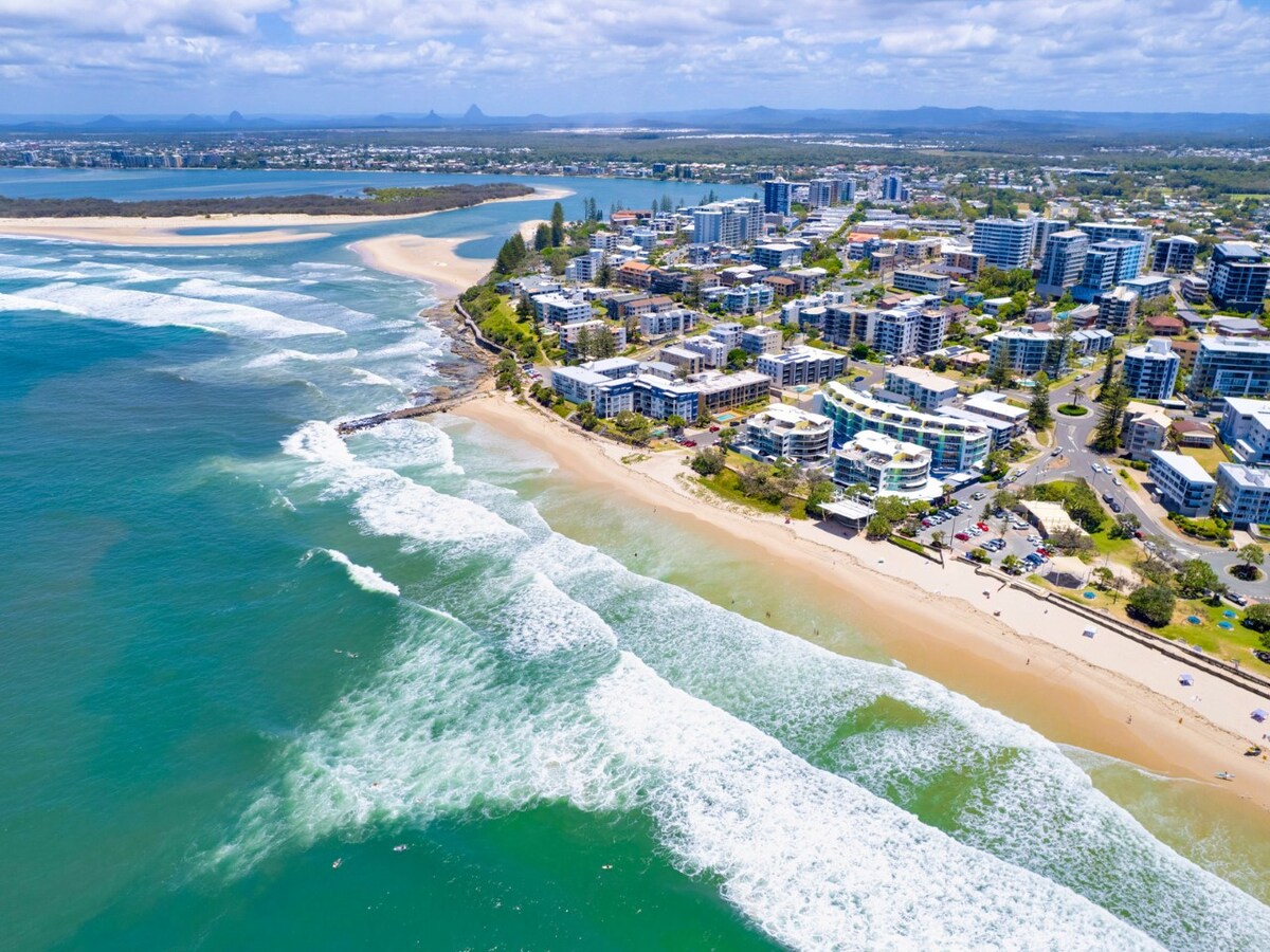 An aerial view showcases a vibrant coastal town with buildings lining the shoreline. The beach is framed by gentle waves lapping against the sand, while lush green landscapes and distant mountains create a serene backdrop.
