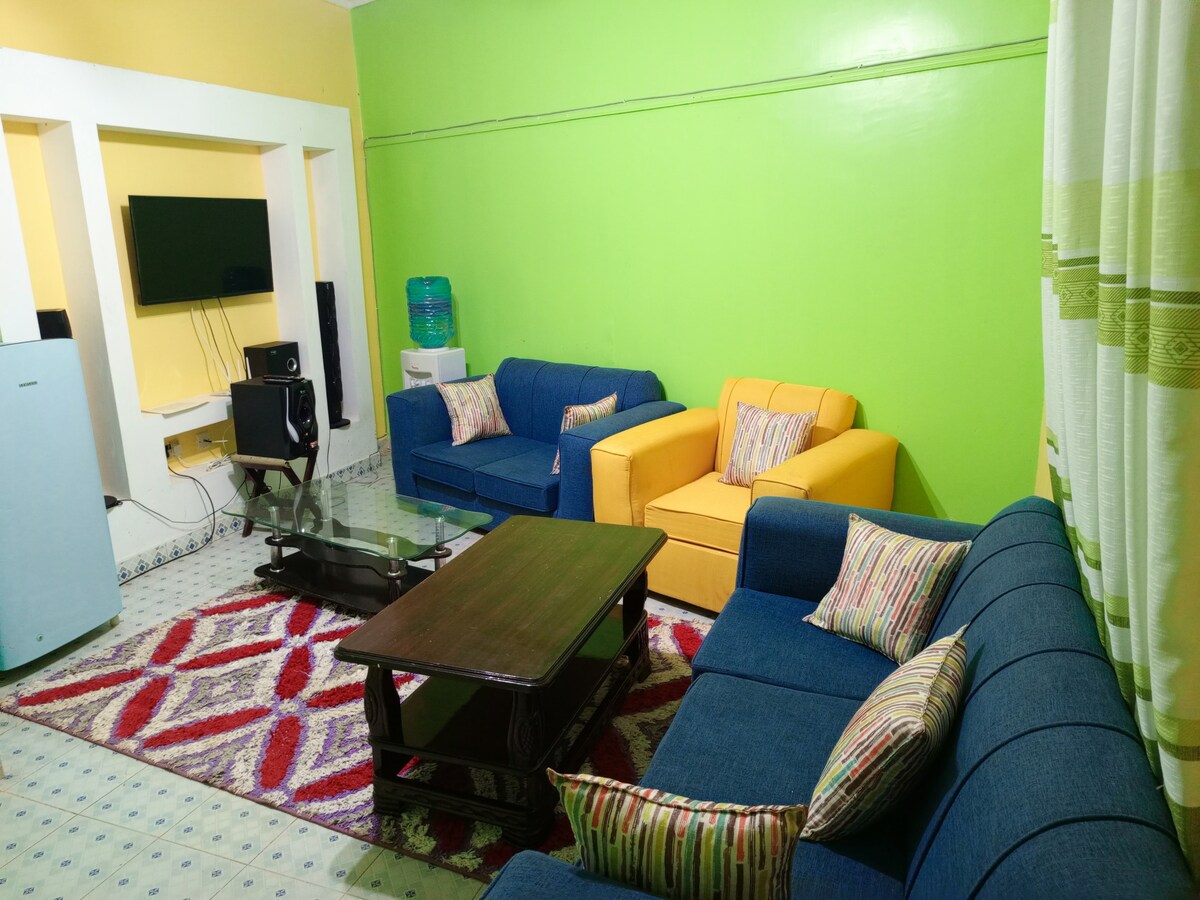 A spacious living area features bright green walls, complemented by a mix of blue and yellow sofas arranged around a glass coffee table. A patterned rug adds warmth to the space, while a television and sound system are positioned against a light-colored wall.