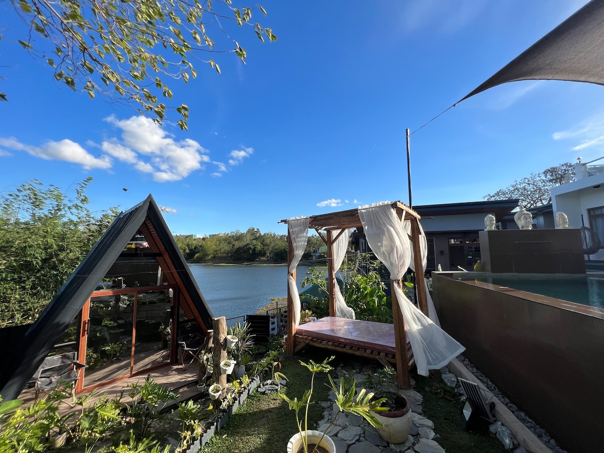 An outdoor pergola bed is situated by the lake, surrounded by lush greenery. A clear blue sky reflects over the water, while a nearby modern structure complements the natural scenery. The tranquil setting promotes relaxation and connection with nature.