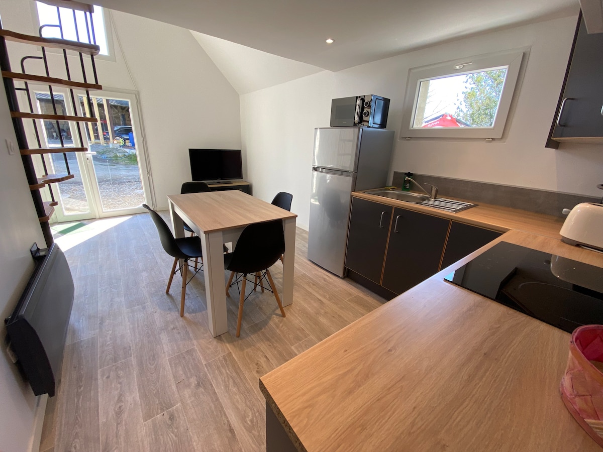 A modern kitchen and dining area are featured, showcasing a wooden table with four black chairs. A compact kitchen with sleek cabinetry, a refrigerator, and a microwave is visible. Natural light pours in through a large window, illuminating the space.