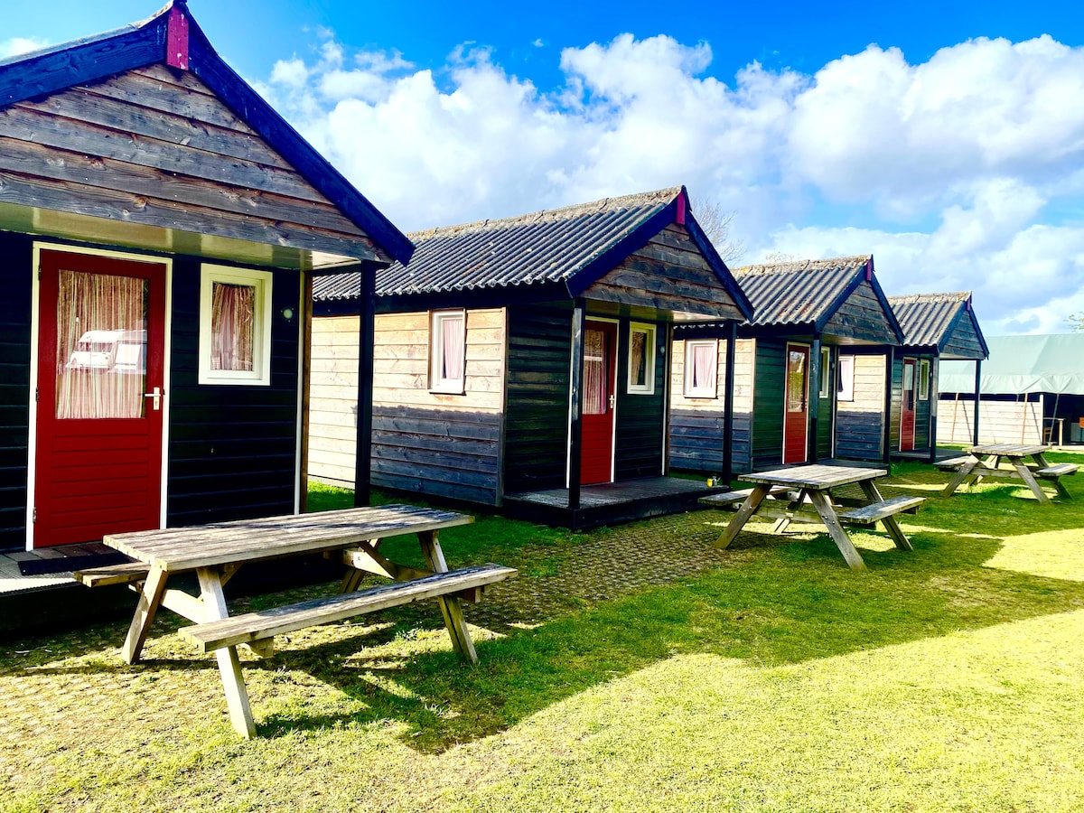 A row of wooden cabins with red doors is positioned on a grassy area. Each cabin features a simple design with a peaked roof. Wooden picnic tables are situated in front of the cabins, providing outdoor seating options under a partly cloudy sky.