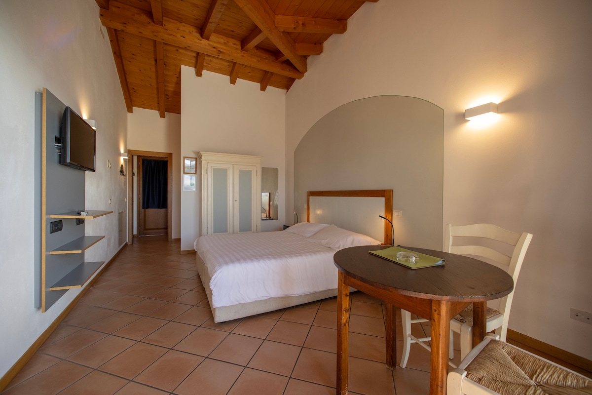 An intimate studio features a spacious bed with crisp white linens, accompanied by a wooden table and chair. A flat-screen TV is mounted on the wall. The room showcases a high wooden ceiling with exposed beams, enhancing a sense of openness and warmth.