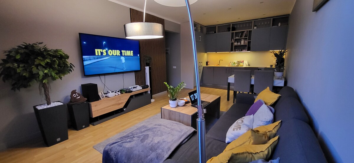 A modern living area features a large television mounted on a wall, displaying vibrant text. A cozy sectional sofa is adorned with decorative pillows, complemented by a wooden coffee table. Natural light fills the space through an open kitchen area, showcasing contemporary cabinetry.