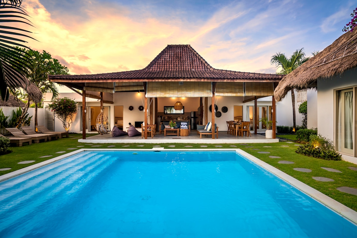 The villa features a spacious outdoor area with a lagoon pool in vibrant turquoise, surrounded by lush grass. The traditional roof and open living space create an inviting atmosphere, while tropical plants and comfortable seating areas enhance the leisure environment.