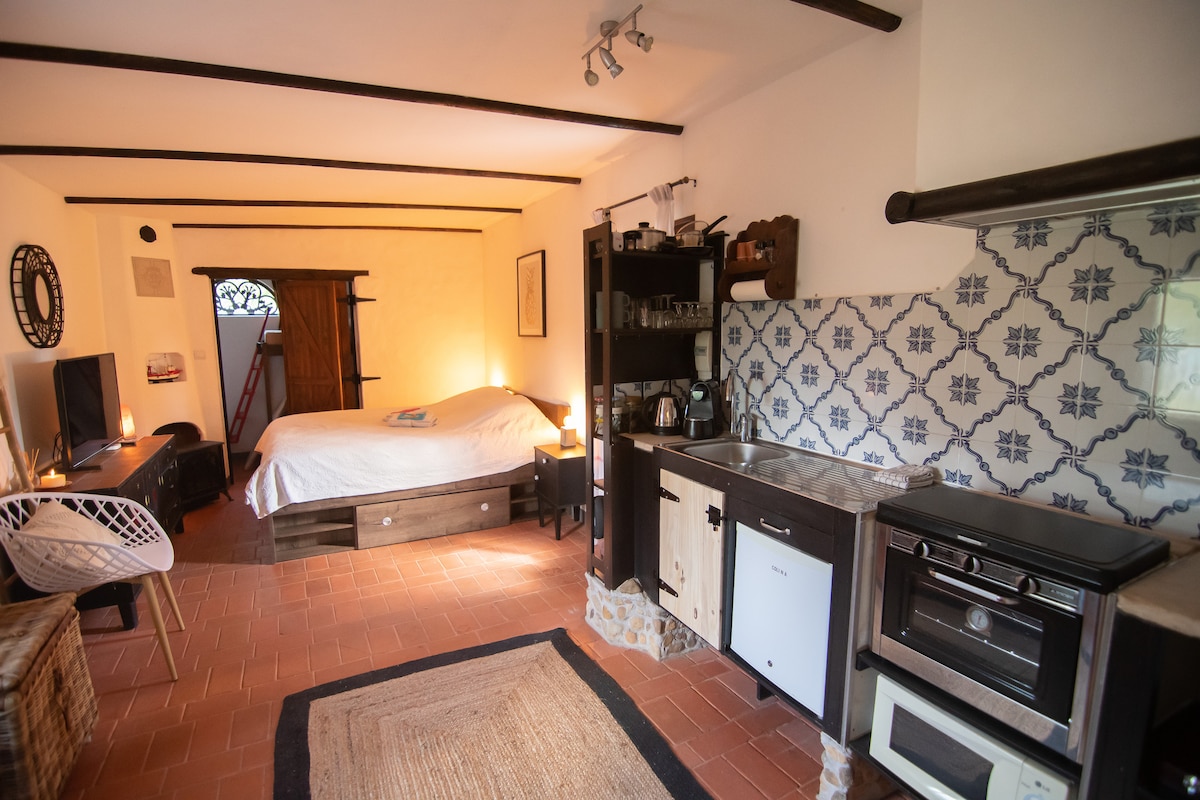A cozy interior is showcased, featuring a spacious bed with neutral bedding and wooden accents. A kitchenette with tiled backsplash and modern appliances is visible, alongside a comfortable seating area. The warm colour palette creates a relaxed atmosphere, complemented by natural light from a nearby window.
