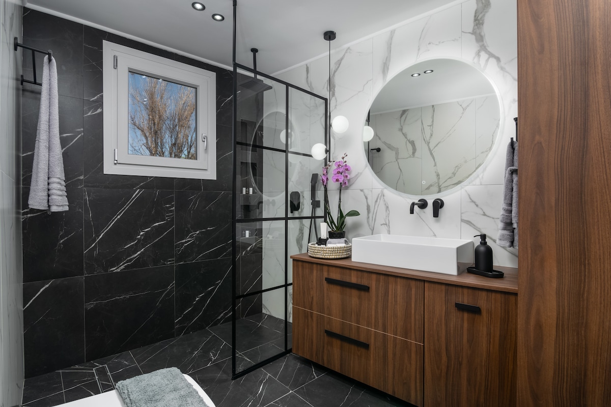 The modern bathroom features dark marble walls and a spacious glass shower. A sleek wooden vanity holds a white sink, accompanied by a round mirror. Natural light enters through a window, and decorative elements include an orchid and neatly arranged towels.