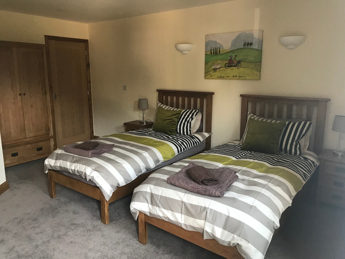Two single beds are positioned in a spacious room, featuring striped bedding in shades of green and beige. Each bed is complemented by a bedside table and lamp. A framed artwork depicting a rural scene hangs above the beds, and wooden wardrobes are visible along the wall.