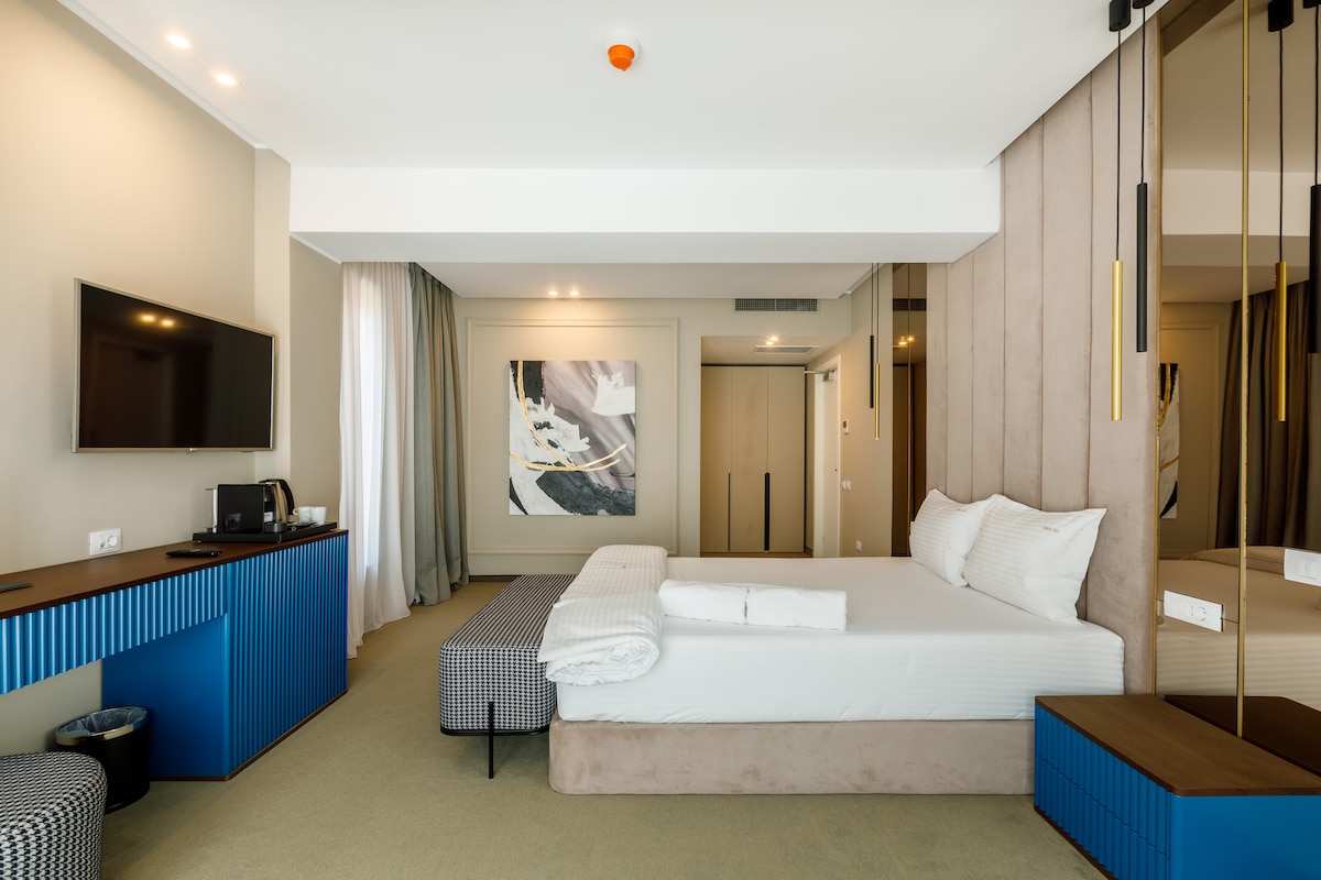 An elegant bedroom features a spacious bed with soft linens, complemented by a blue accent wall and sleek furniture. A large flat-screen TV is mounted on the wall, and a coffee station is available. Natural light filters through sheer curtains, enhancing the space's openness.