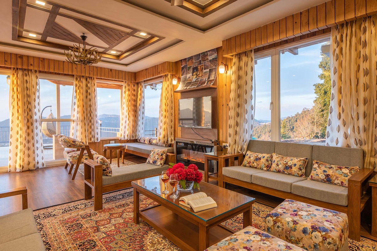 An inviting living area showcases elegant wooden furnishings and large windows overlooking scenic vistas. Floral-patterned sofas and an area rug add warmth to the space, while a coffee table displays fresh flowers and a book. Soft lighting enhances the comfortable atmosphere.