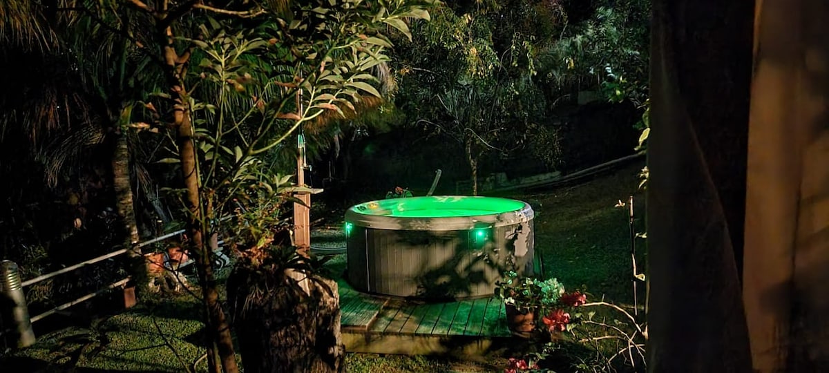 A circular jacuzzi is positioned on a wooden deck surrounded by lush greenery. Soft green lights illuminate the water, creating a serene atmosphere in the evening setting. Potted plants add to the natural ambiance, while the surrounding foliage enhances privacy.