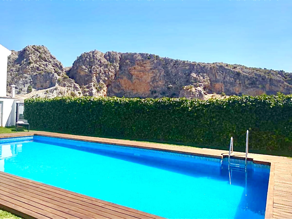 A clear swimming pool occupies the foreground, reflecting the blue sky above. Surrounding greenery adds privacy, with rugged natural stone formations visible in the background. A wooden deck frames the pool area, enhancing the tranquil outdoor setting.