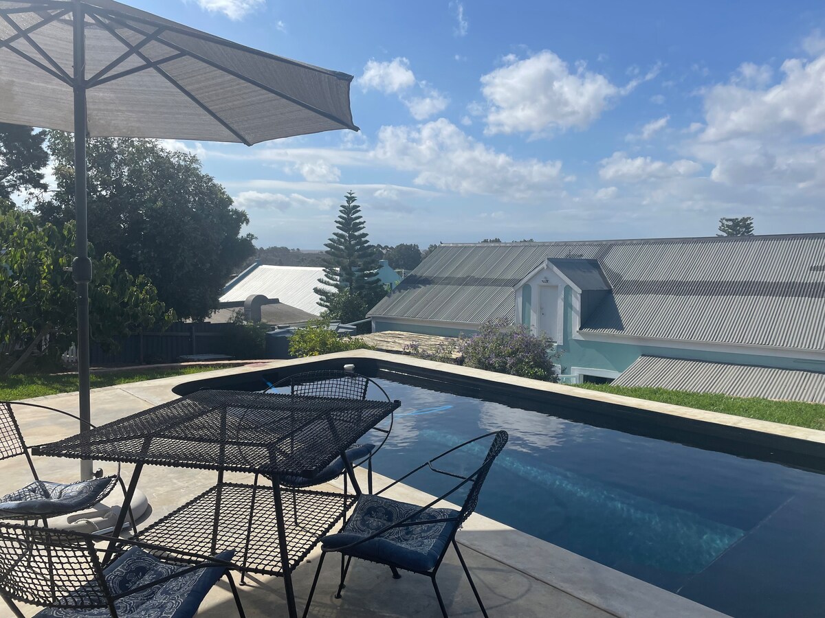 An outdoor seating area features a round table surrounded by black metal chairs, offering a view of the swimming pool. An umbrella provides shade, and a clear sky with scattered clouds enhances the tranquil atmosphere of the garden area.