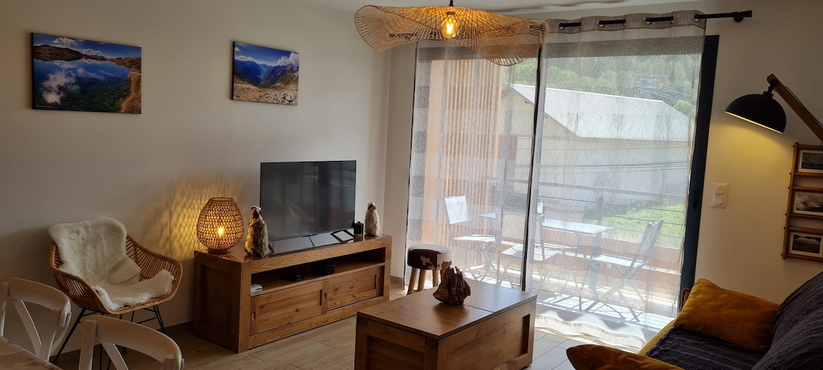 The spacious living area features a modern flat-screen TV placed on a rustic wooden console. Natural light filters through sheer curtains, illuminating a cozy seating arrangement and a coffee table, while a balcony with outdoor seating can be viewed outside.