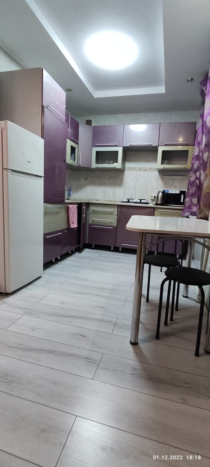 A compact kitchen is presented with purple cabinetry and modern appliances. White countertops are complemented by a neutral backsplash. A small dining table is set with black chairs. Natural light is provided through a ceiling fixture, creating a bright atmosphere.