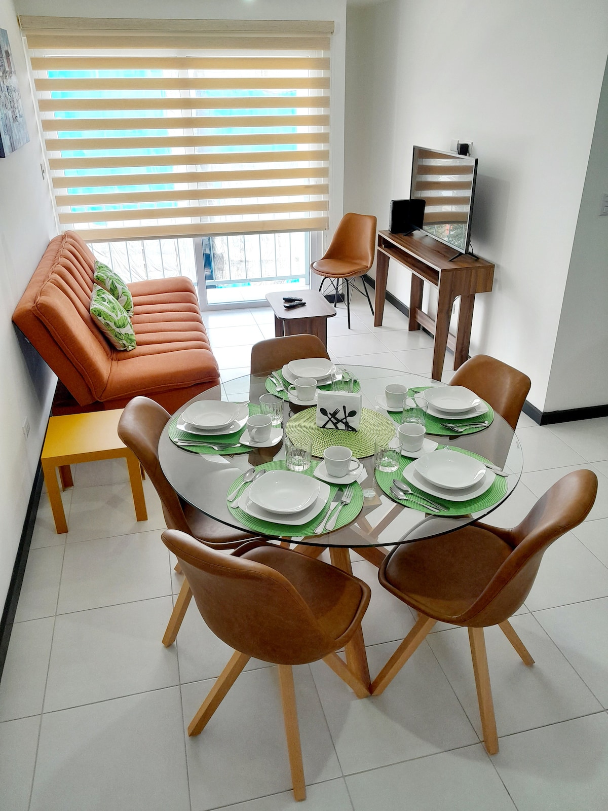 A modern dining area features a round glass table set with dishes, cutlery, and placemats. Brown leather chairs surround the table. To one side, a plush orange sofa provides additional seating, while a TV stand and chair complete the space, and natural light filters through window shades.