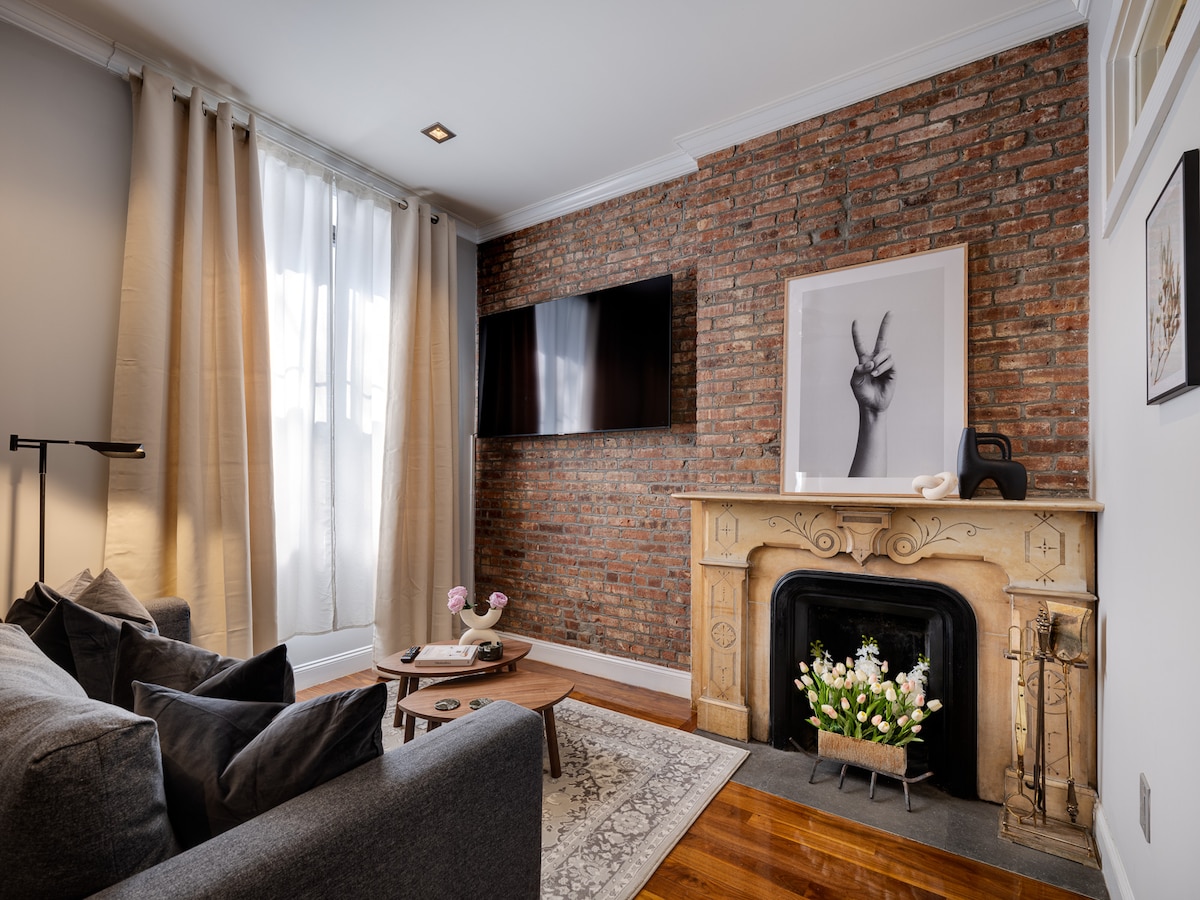 A cozy living area is featured with a dark gray sofa and a decorative coffee table. A framed piece of art hangs above a decorative fireplace, complemented by a bouquet of flowers. Natural light shines through softly draped curtains near the exposed brick wall.