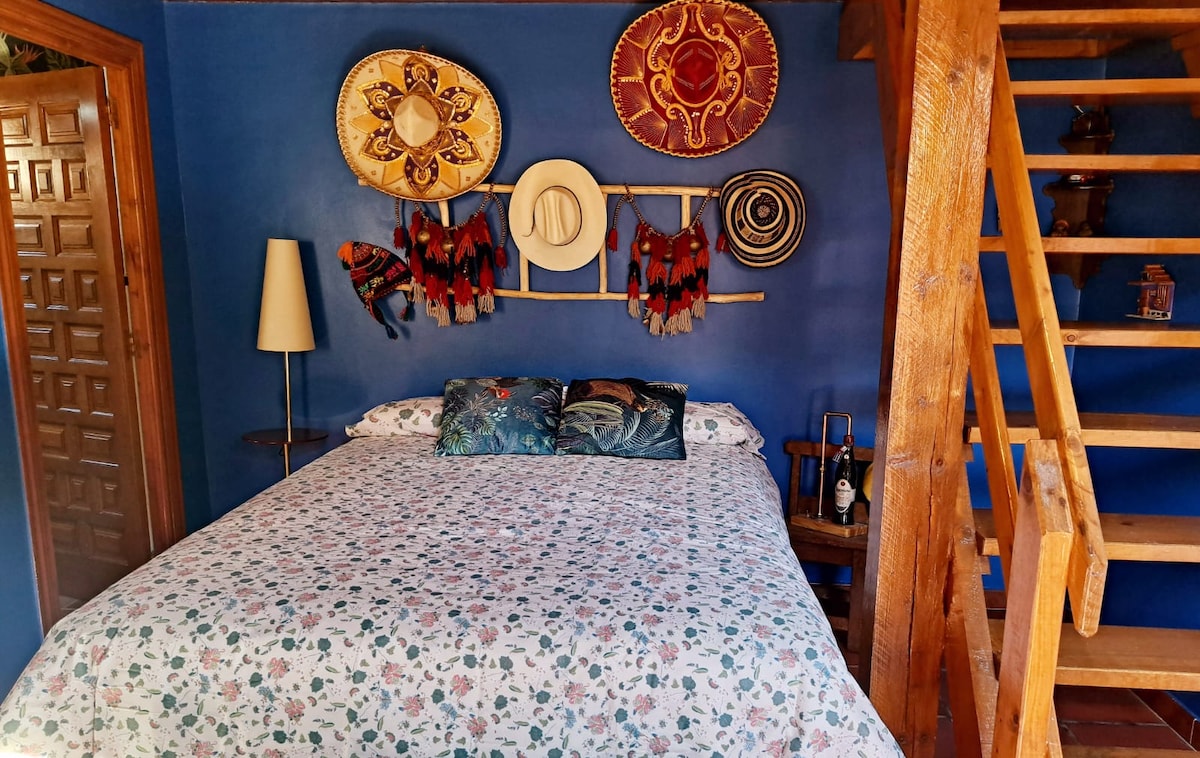 A sleeping area showcases a spacious bed adorned with a floral-patterned bedspread. The walls are painted in a vibrant blue, complemented by decorative hats and textiles arranged above the bed. A wooden staircase leads to the upper level, enhancing the rustic charm of the space.