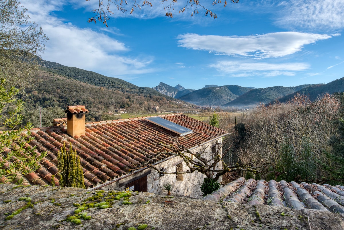 A traditional rural house with a tiled roof is framed by lush green hills and mountains in the background. The landscape features a clear blue sky with scattered clouds, and a serene valley is visible, enhancing the tranquil setting.
