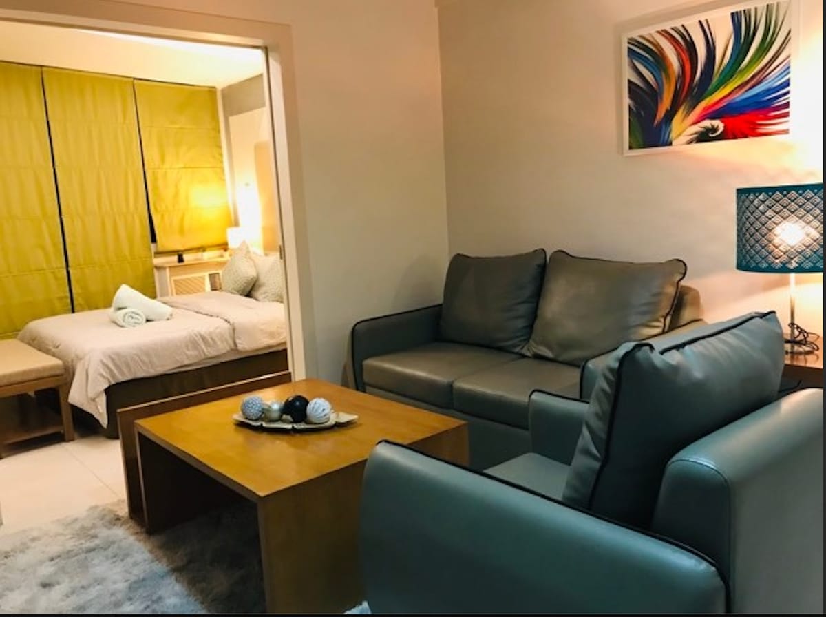 A cozy living area includes a comfortable gray sofa and an armchair, facing a wooden coffee table. A colorful wall art piece complements the space. In the background, an illuminated bedroom can be seen, featuring a large bed with neatly arranged linens and a calming color palette.