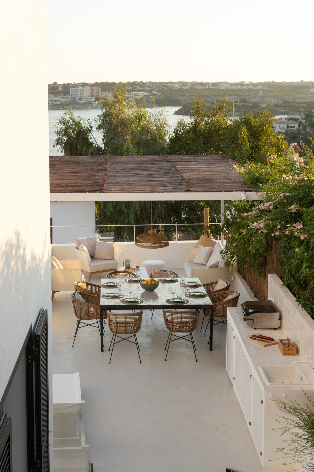 An outdoor dining area is set on a spacious terrace, featuring a large table surrounded by stylish chairs. The space includes a sofa arrangement with plush cushions. A view of the water is visible in the background, framed by greenery and soft rays of sunlight.