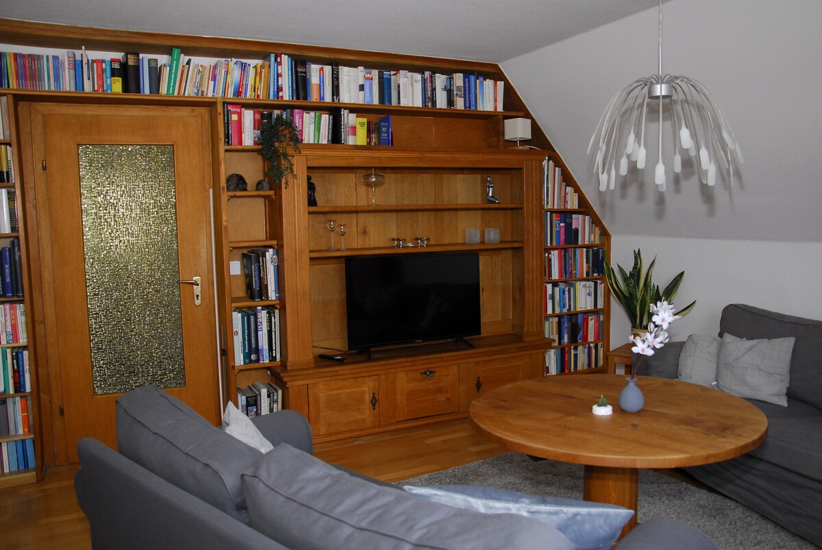 A living area is presented with wooden shelving units filled with books. A round wooden table is positioned at the center, accompanied by gray sofas. A television is mounted on the wall beneath a decorative wooden shelf, enhancing the cozy environment.