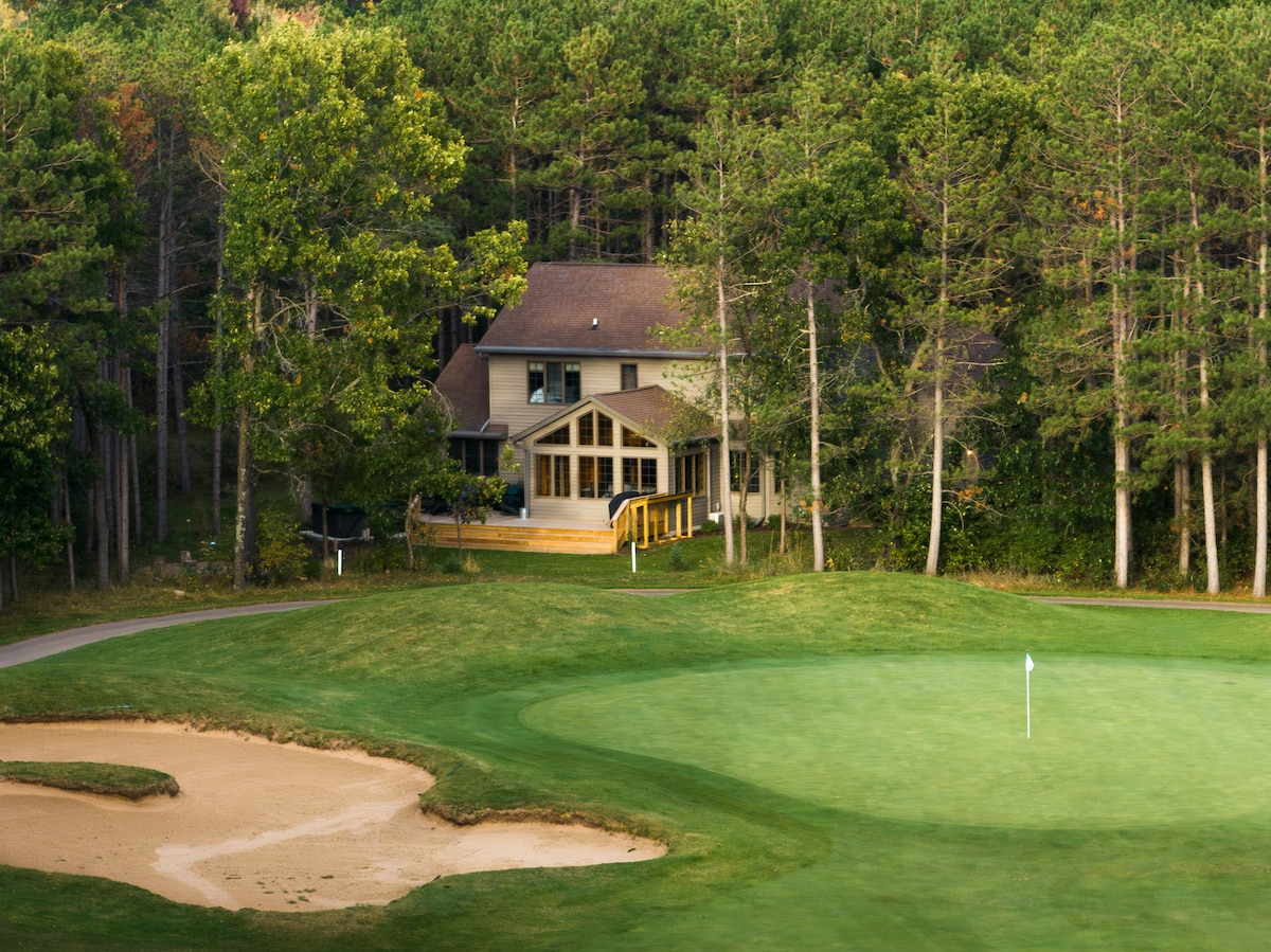 The exterior view depicts a two-story home nestled among tall trees, providing a sense of seclusion. A manicured golf course is visible in the foreground, featuring a sandy bunker and a green with a pin. A pathway leads to the home's entrance.