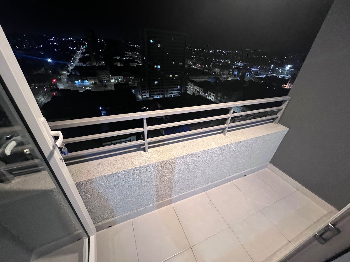 A balcony area is shown, featuring a low railing and tile flooring. The city skyline is visible at night, with street lights and buildings illuminated. A feeling of space is conveyed, inviting guests to enjoy the urban view.