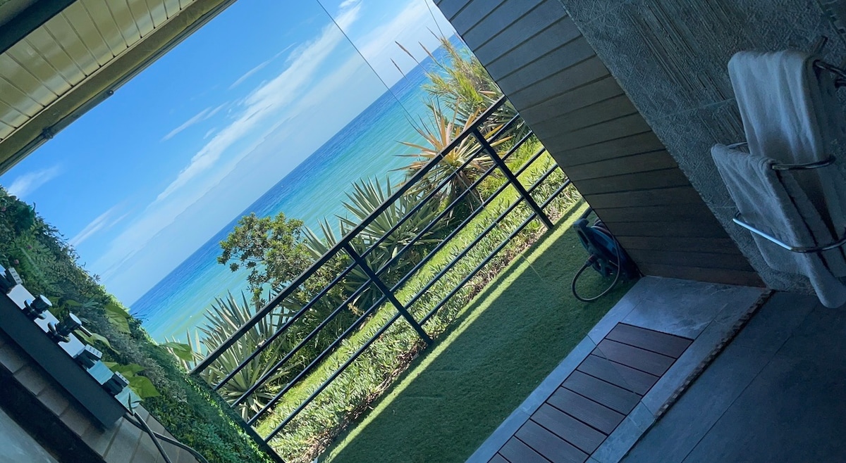 A serene outdoor view is presented, showcasing lush greenery and a vibrant blue sea. The image captures a seamless transition from the indoors to the outdoor space through large glass sliding doors, enhancing the feeling of openness and connectivity with nature.