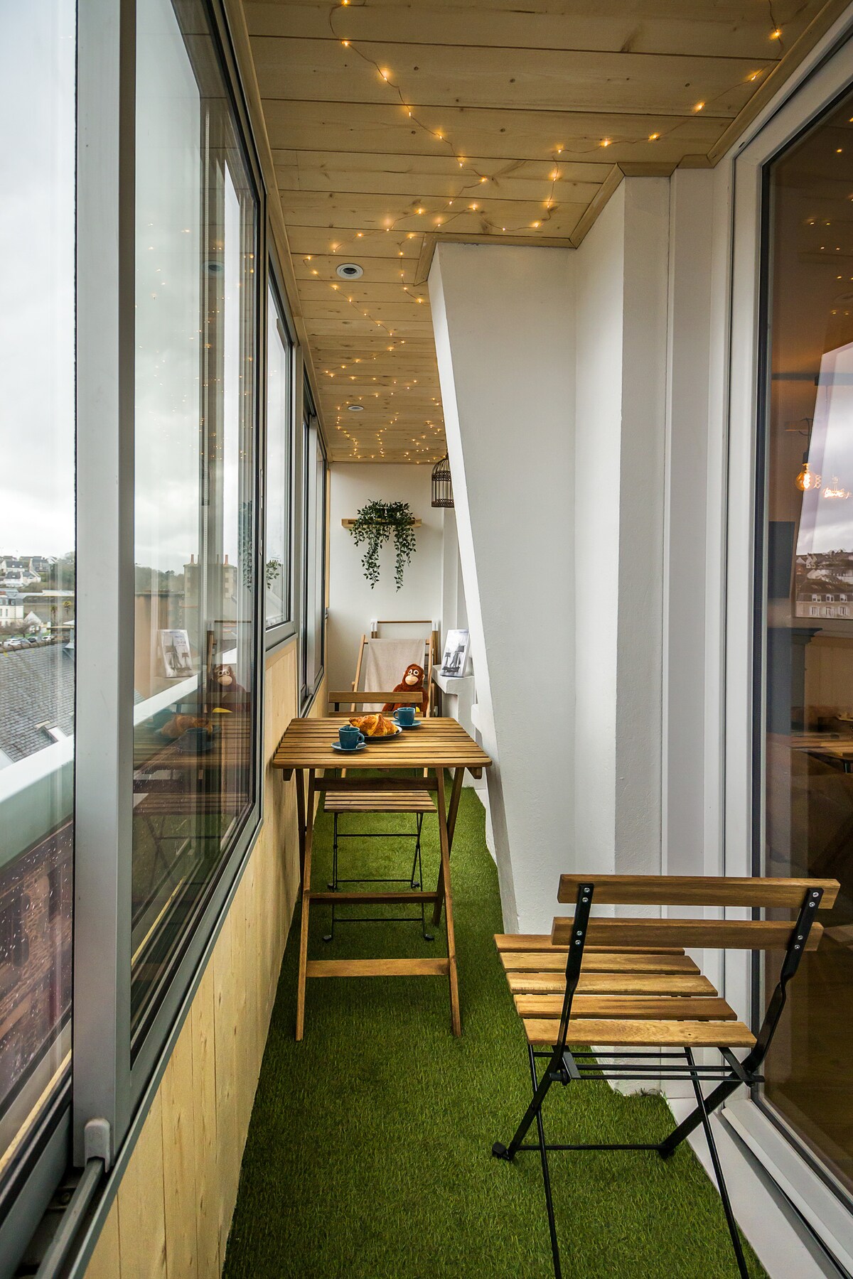 A closed balcony features two wooden chairs and a small table, inviting outdoor dining. Decorative fairy lights are strung across the ceiling, and greenery decorates the wall, creating a cozy atmosphere. The floor is covered with artificial grass, enhancing the inviting setting.