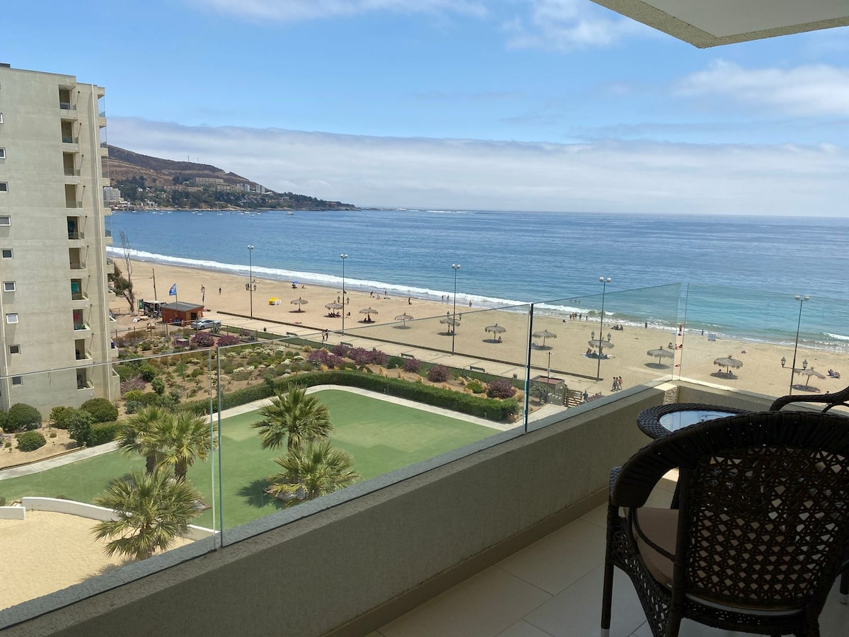 A balcony view showcases a sandy beach and the ocean. Palm trees and beach umbrellas are visible along the shoreline, while a grassy area is seen below. The sky is bright and clear, enhancing the scene of a serene beach atmosphere.