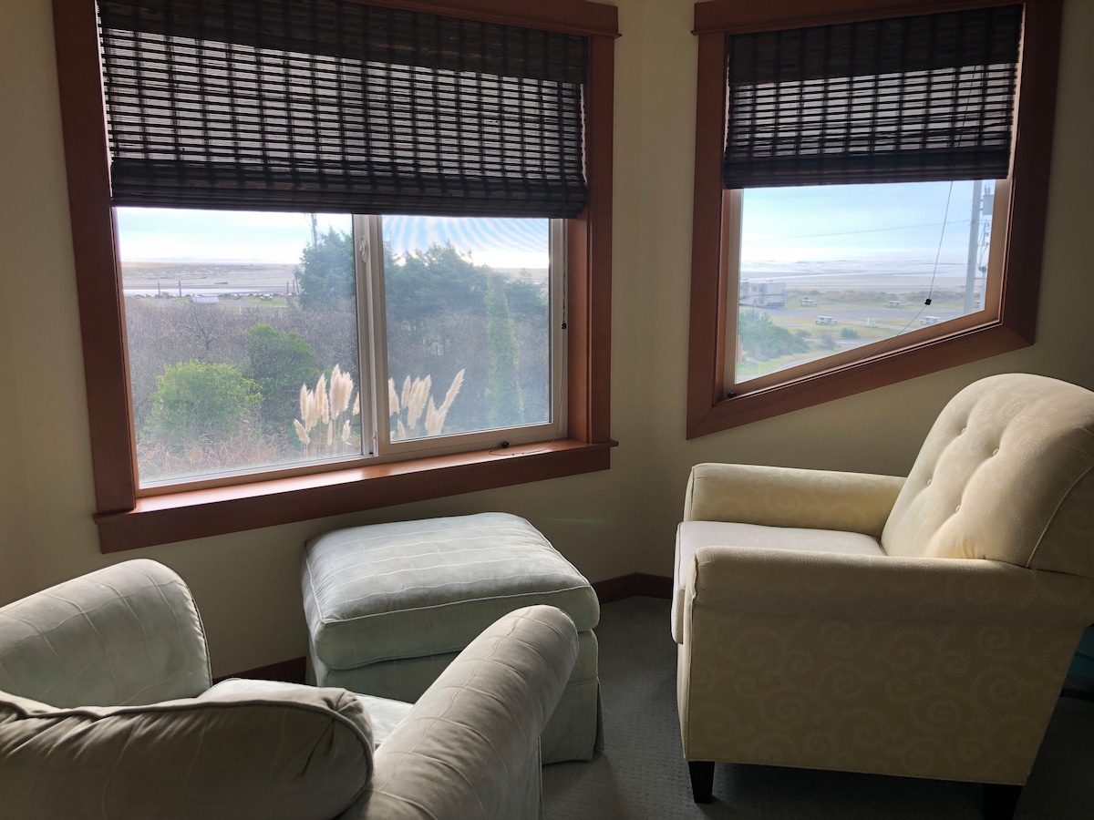 A cozy corner nook offers views of the coastal landscape through large windows. Two light-colored armchairs provide comfortable seating, accompanied by a soft ottoman. Natural light fills the space, while blinds add privacy without obstructing the scenery.