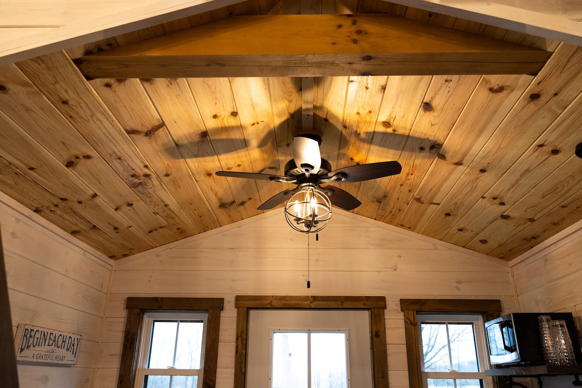 The interior features a high, sloped wooden ceiling with exposed beams, creating a spacious feel. A ceiling fan with a light fixture is centrally located, complemented by two framed windows on either side that allow natural light to enter the room.