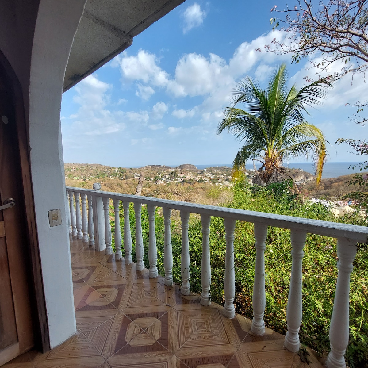 A balcony overlooks a lush landscape featuring hills and scattered white buildings. A tall palm tree stands at the edge of the view, under a partly cloudy sky. The ground is tiled with decorative patterns, enhancing the outdoor space.