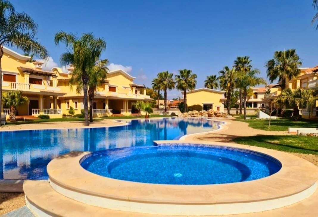 A spacious outdoor area features a large swimming pool with a separate circular hot tub. Lush palm trees surround the pool, while sun loungers are placed by the water. The backdrop includes elegant residential buildings set amidst well-maintained gardens.