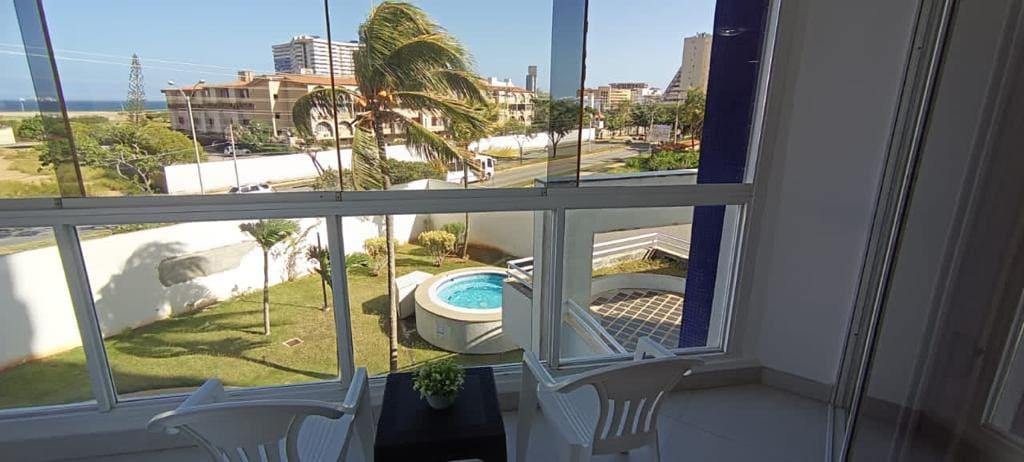 A view captured from a window displays a well-maintained outdoor area featuring a hot tub surrounded by grass. The scene includes palm trees swaying gently in the breeze and a glimpse of the ocean in the background.