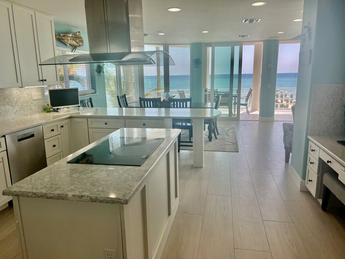 A modern kitchen with light cabinetry and a large island features a stovetop and ample counter space. Floor-to-ceiling windows provide views of the ocean and natural light. A dining area with wooden chairs is positioned nearby, creating a seamless connection between cooking and dining.